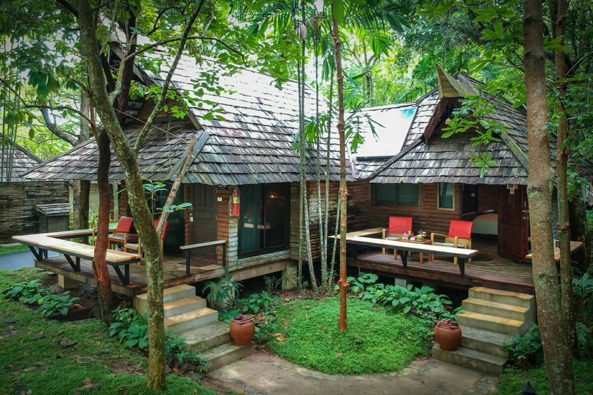 A secluded resort with wooden cabins surrounded by lush greenery. Each cabin features a deck with seating and a table.