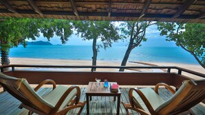 Terraza de madera con dos sillas y una mesa, con vista a una playa de aguas azules y montañas distantes.