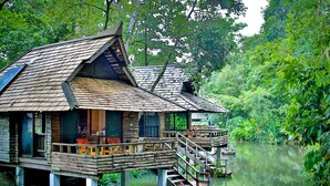 Una casa flotante de madera con terraza y escalera, navegando sobre aguas tranquilas rodeada de exuberante vegetación.