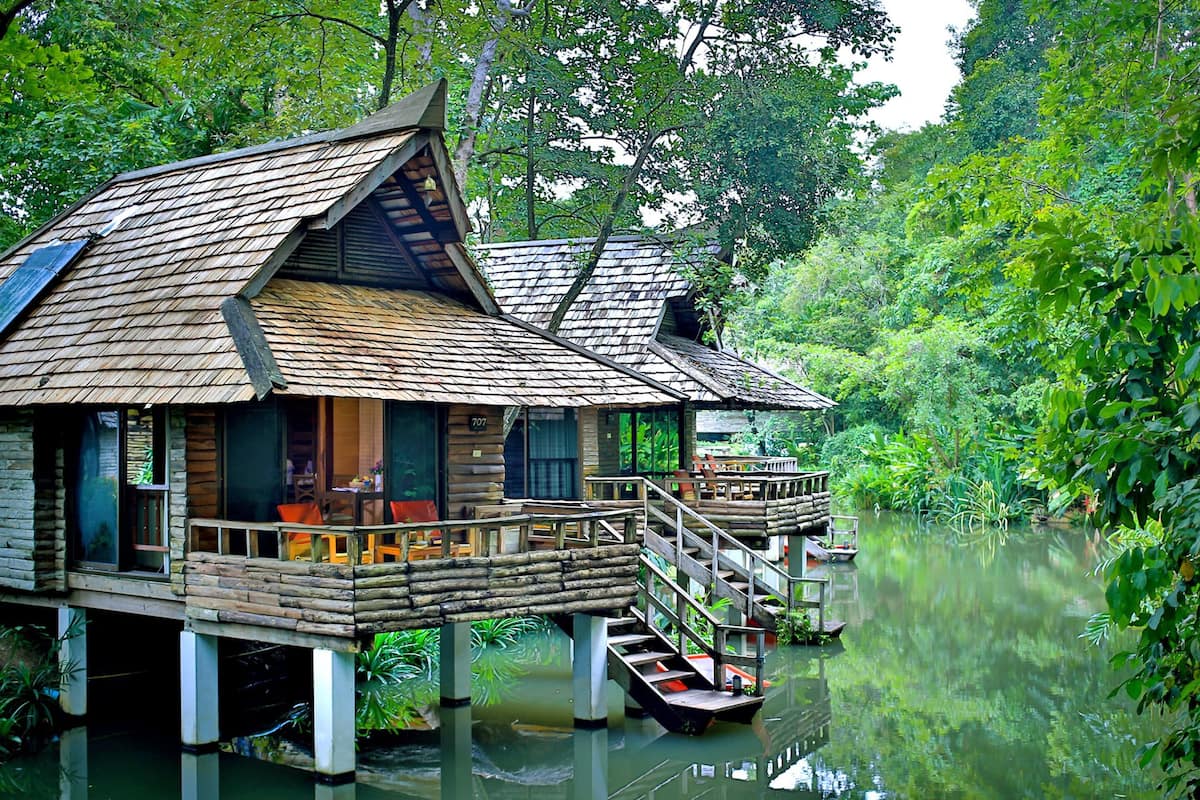 A wooden houseboat with a deck and a staircase, floating on calm water surrounded by lush greenery.