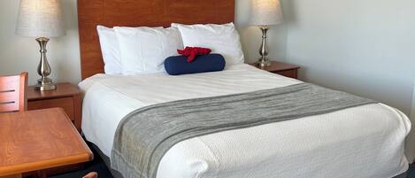 Una cama bien tendida con sábanas blancas, cabecera de madera y una manta gris. Hay dos lámparas de noche, una mesita de noche de madera y un cuadro enmarcado en la pared.