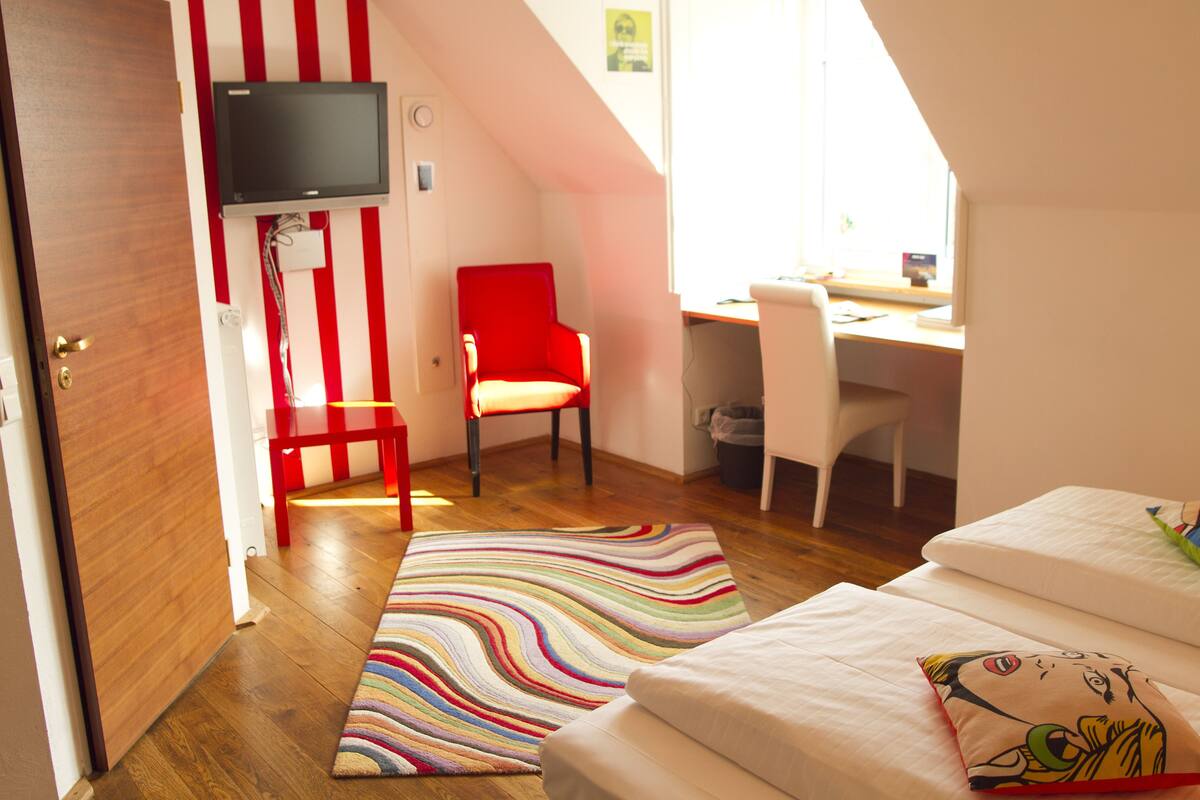 Une chambre à coucher avec un lit, un tapis rayé aux couleurs vives, une chaise rouge, un bureau et un téléviseur fixé au mur.