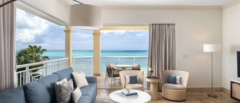 A modern living room with a sofa, armchairs, a coffee table, and a TV. It features a balcony with a view of the ocean.