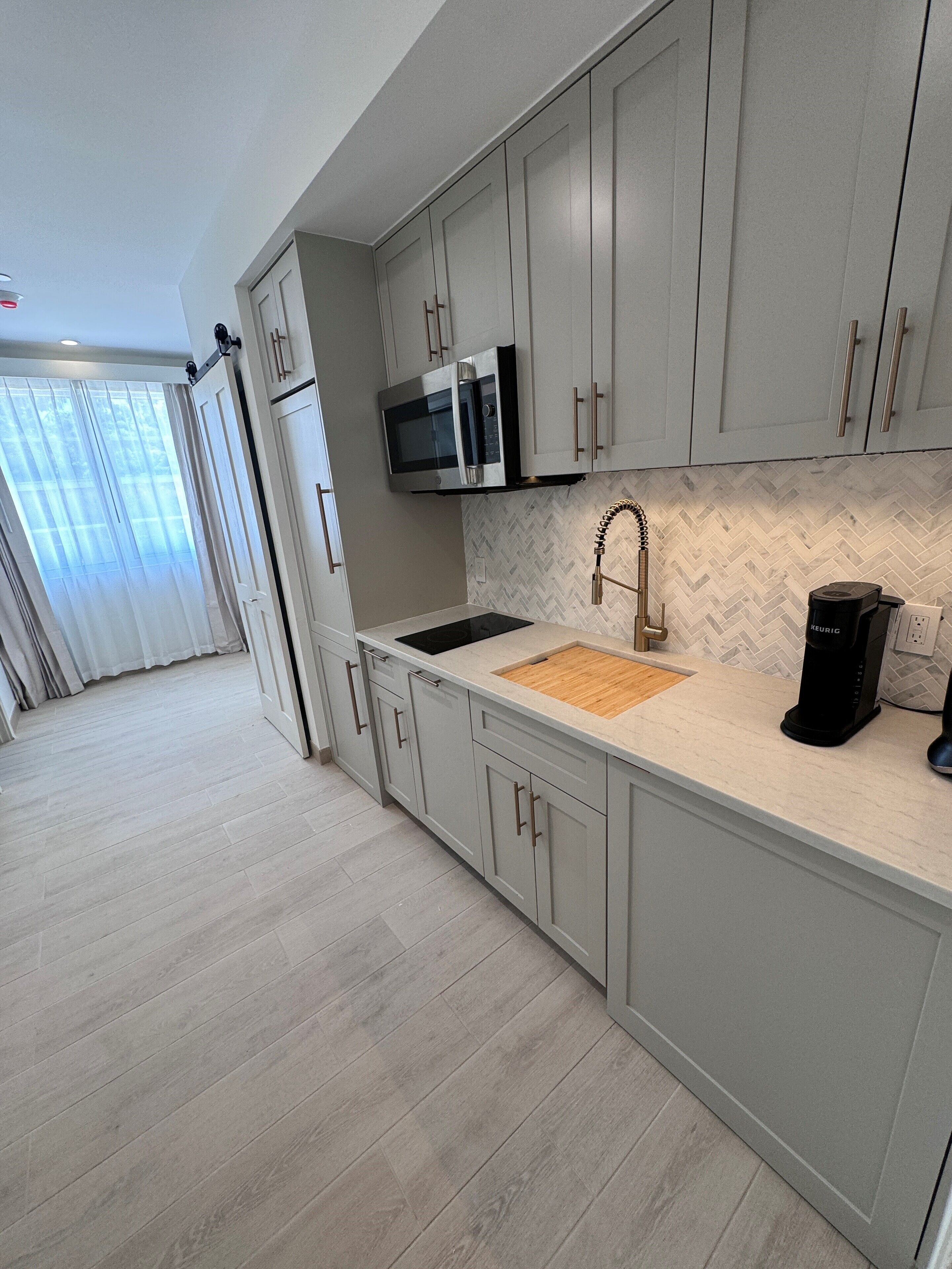 A modern kitchen with grey cabinets, a built-in microwave, a sink with a wooden cutting board, and a coffee machine.