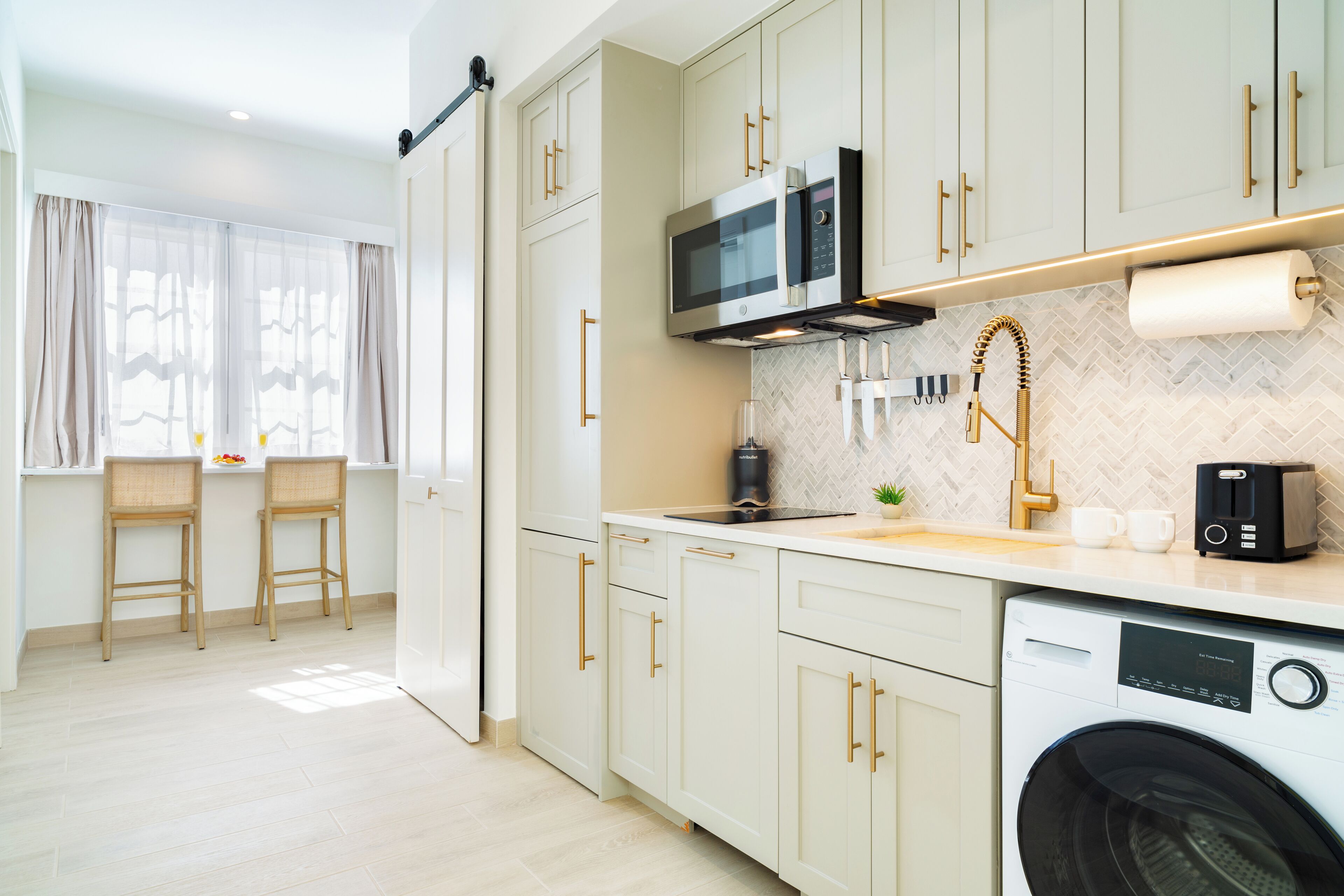 A modern kitchen with a microwave, coffee maker, and washing machine. There are two bar stools and a window with sheer curtains.
