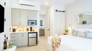 A modern hotel room with a kitchenette, a bed with floral bedding, a large mirror, and a bedside table with a lamp.