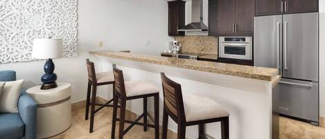 A modern kitchen with dark wood cabinets, a stainless steel refrigerator, a granite countertop, and a bar area with two bar stools.