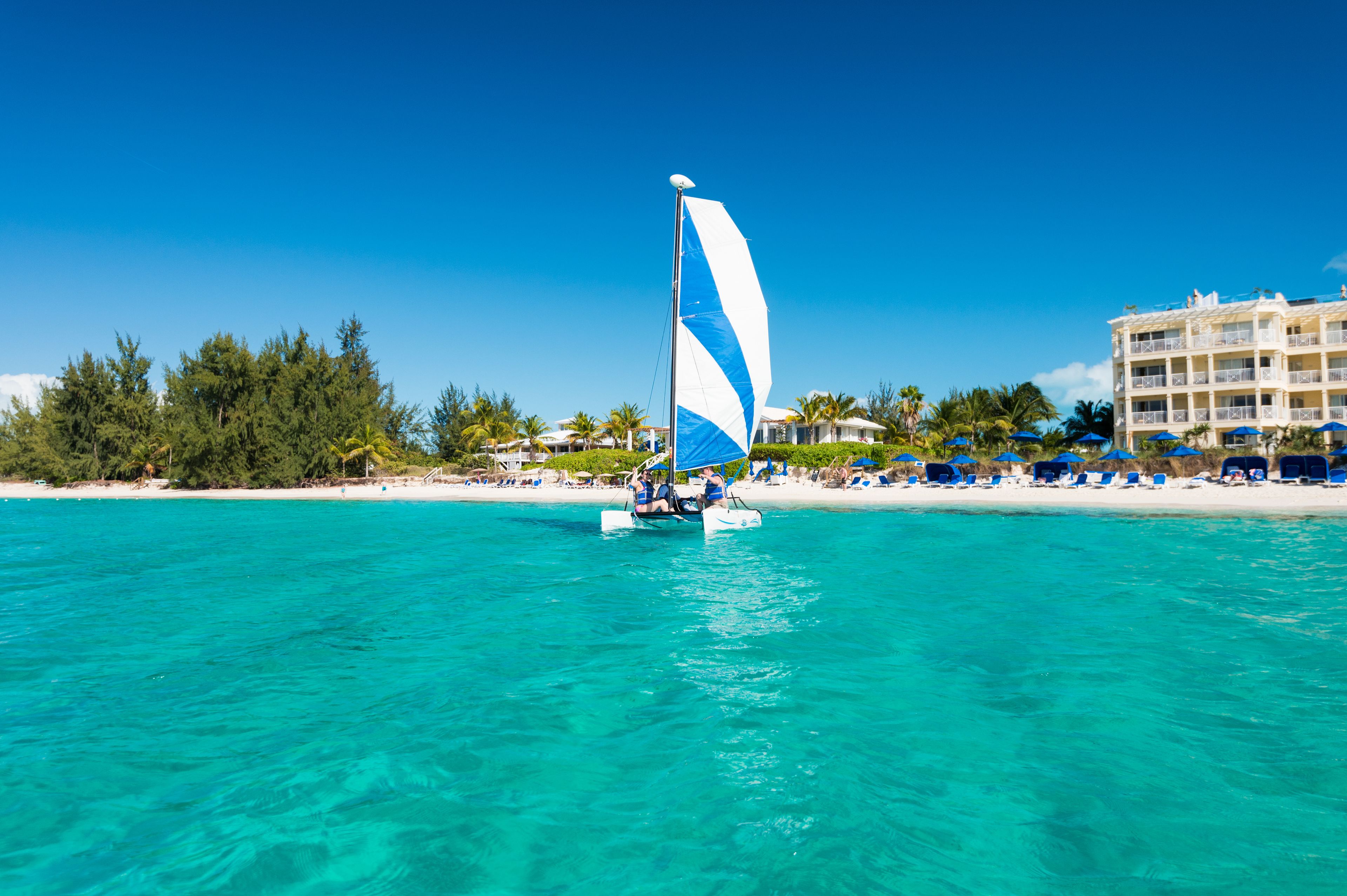 Un velero sobre aguas turquesas, con una playa y edificios de un complejo turístico al fondo.