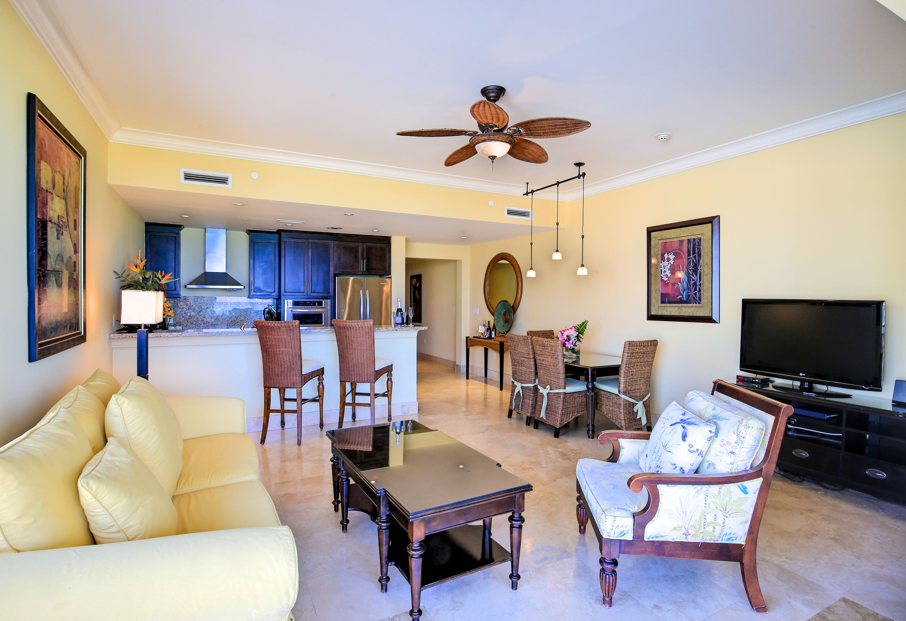 A spacious living room with a sofa, coffee table, and chairs, a dining area with a table and chairs, a kitchen with cabinets and a refrigerator, and a ceiling fan.