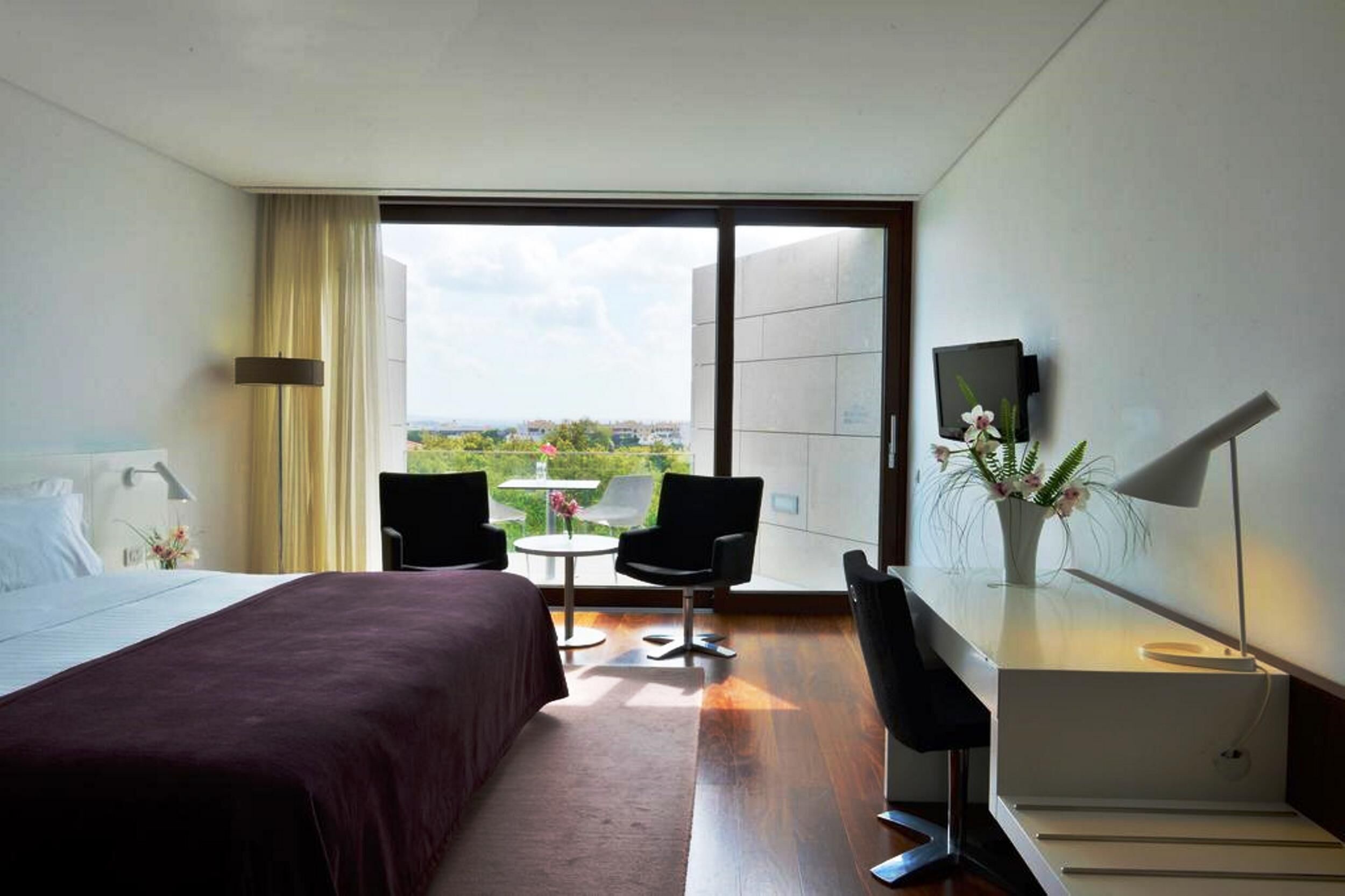 Habitación de hotel con cama, dos sillones, una mesita, televisor y vistas a zonas verdes a través de amplios ventanales.