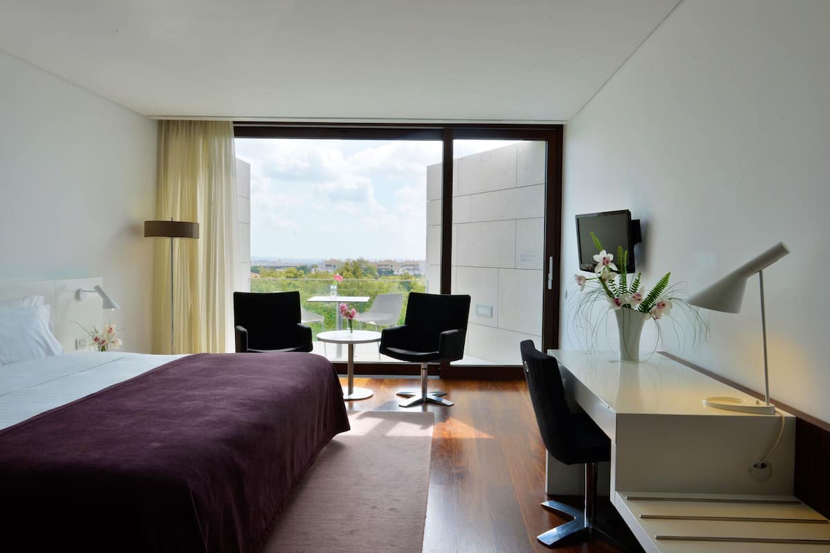 Habitación de hotel con cama, escritorio, silla, televisor y vistas al exterior.