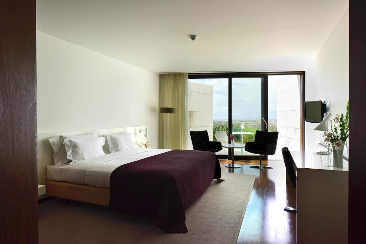 Habitación de hotel con cama, escritorio, dos sillas, televisor y ventana grande con vistas a la ciudad.