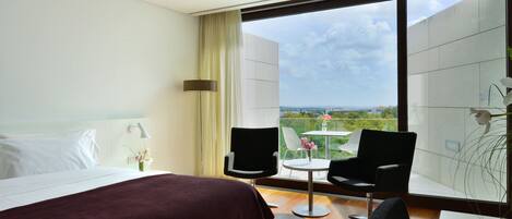 A hotel room with a large bed, two chairs, a small table, and a view of the city.