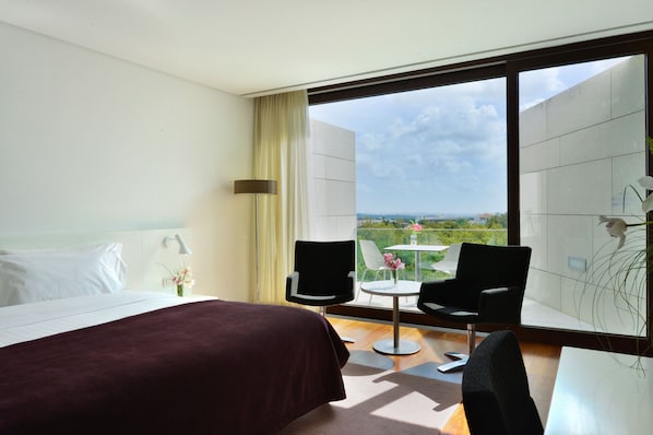 A hotel room with a large bed, two chairs, a small table, and a view of the city.