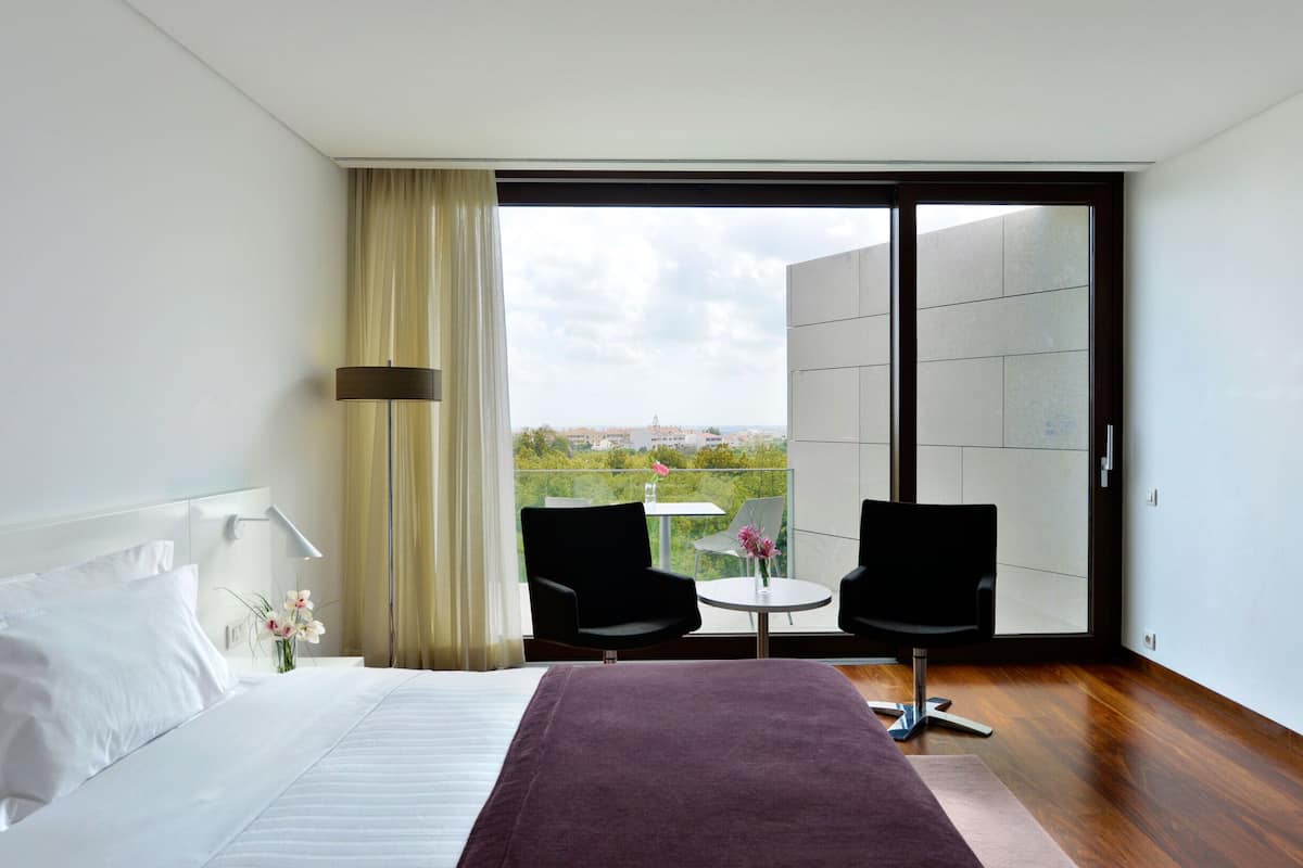Habitación de hotel moderna con una cama grande, dos sillas, una mesita y vistas a través de un ventanal corredero.
