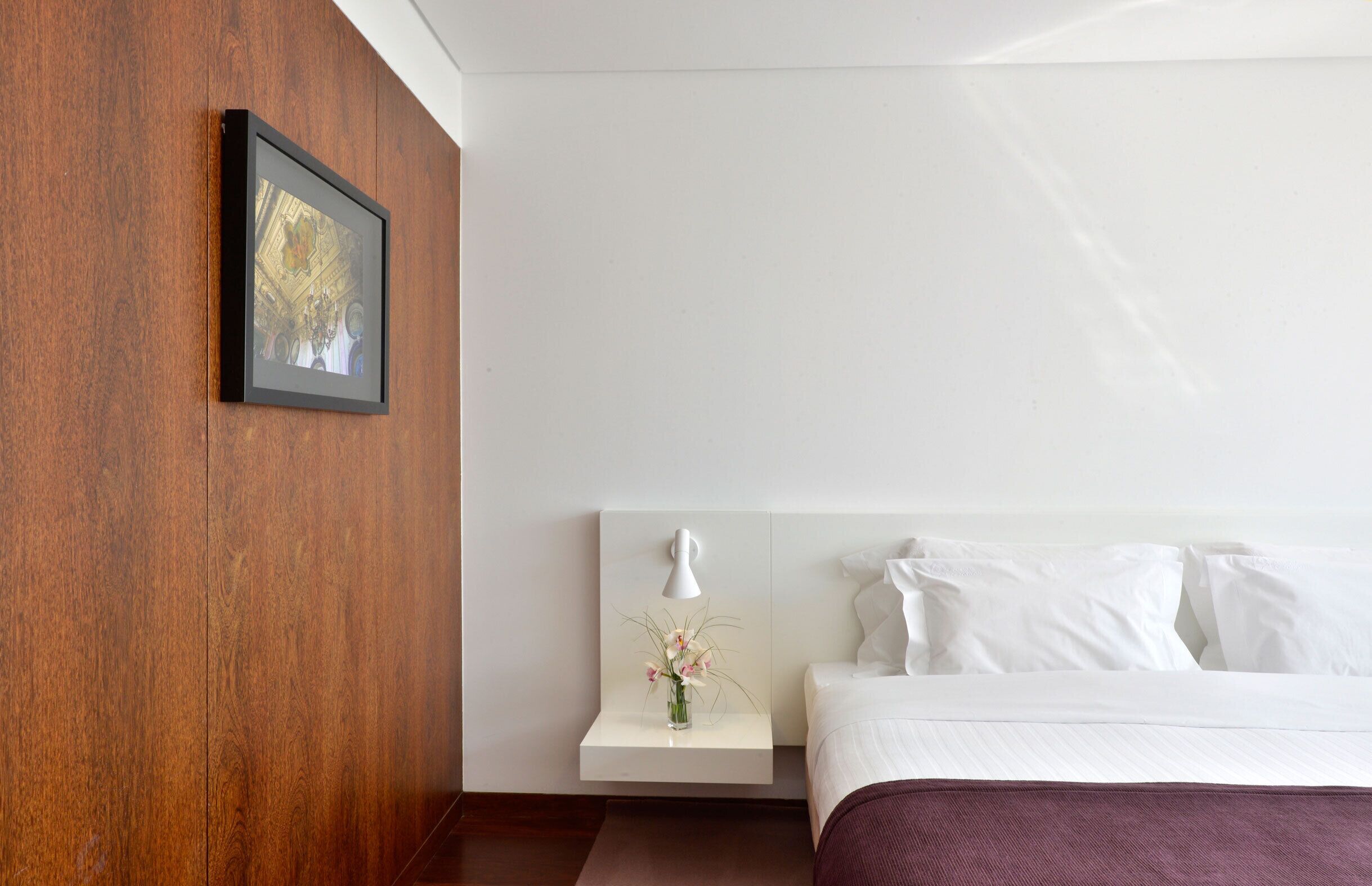 Una habitación de hotel con una pared de madera decorativa, un cuadro enmarcado, una mesita de noche con un jarrón de flores y una cama con ropa blanca.