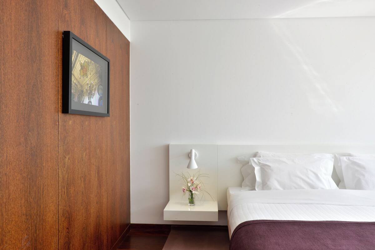 Una habitación de hotel con una pared de acento de madera, un cuadro enmarcado, una mesita de noche con un florero y una cama con ropa de cama blanca.