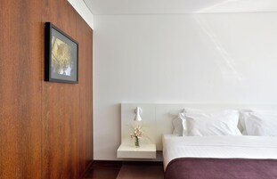 Una habitación de hotel con una pared de acento de madera, un cuadro enmarcado, una mesita de noche con un florero y una cama con ropa de cama blanca.