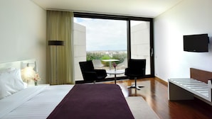 Una habitación de hotel moderna con una cama grande, un televisor montado en la pared, un balcón con mesa y sillas, y vista a la ciudad.