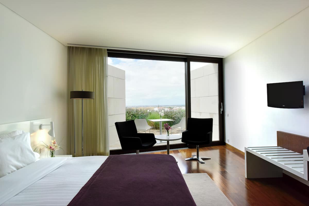 Una habitación de hotel moderna con una cama grande, un televisor montado en la pared, un balcón con mesa y sillas, y vista a la ciudad.