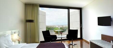 Een moderne hotelkamer met een groot bed, een aan de muur bevestigde televisie, een balkon met een tafel en stoelen, en uitzicht op de stad.