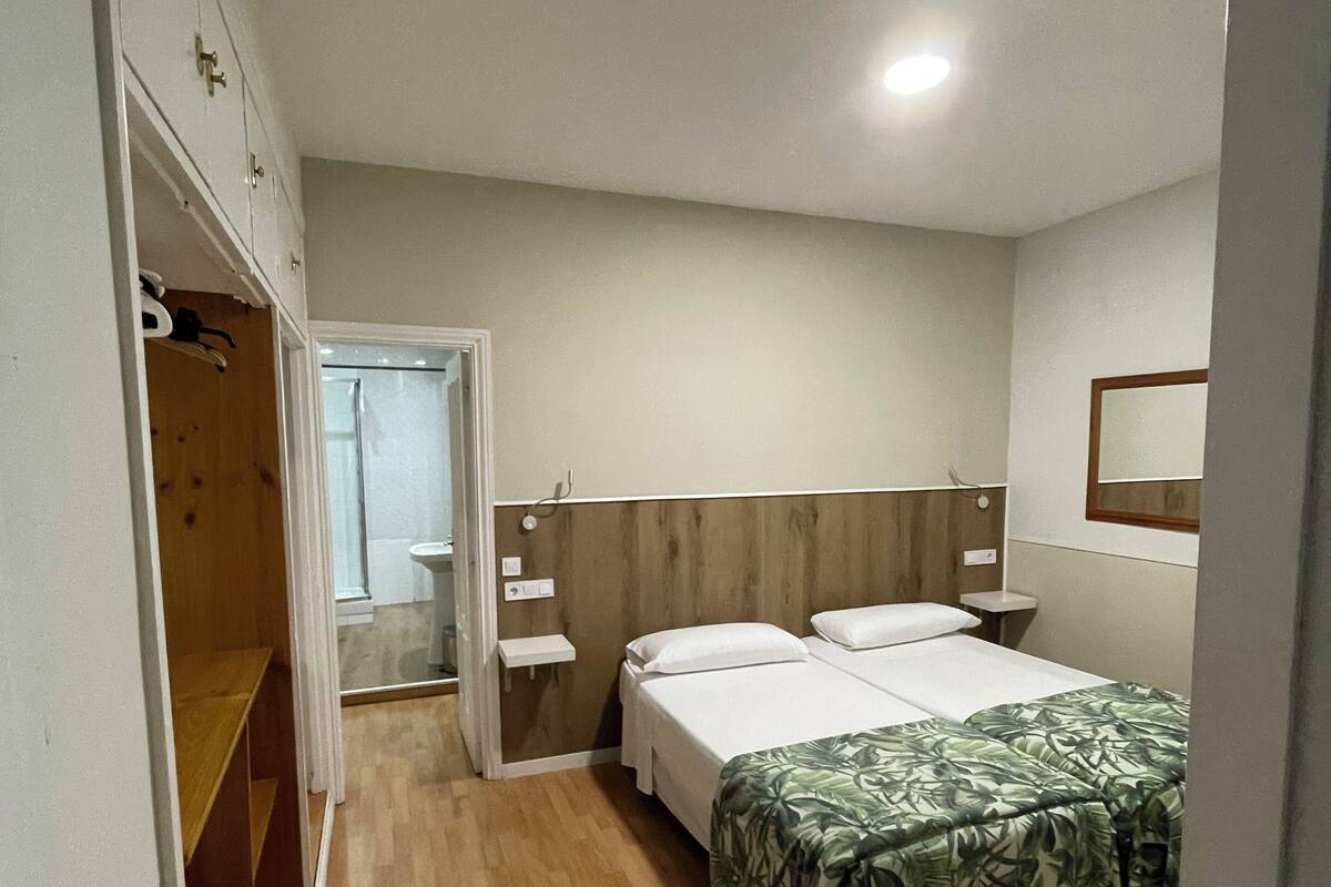 Un dormitorio con piso de madera, una cama con cubrecama estampado de hojas verdes, una mesita de noche y un buró.