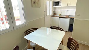 A compact kitchen with white cabinets, a microwave, and a small refrigerator. There is a dining table with four chairs and a window with blinds. - Apartamentos Mayor Centro (Madrid)