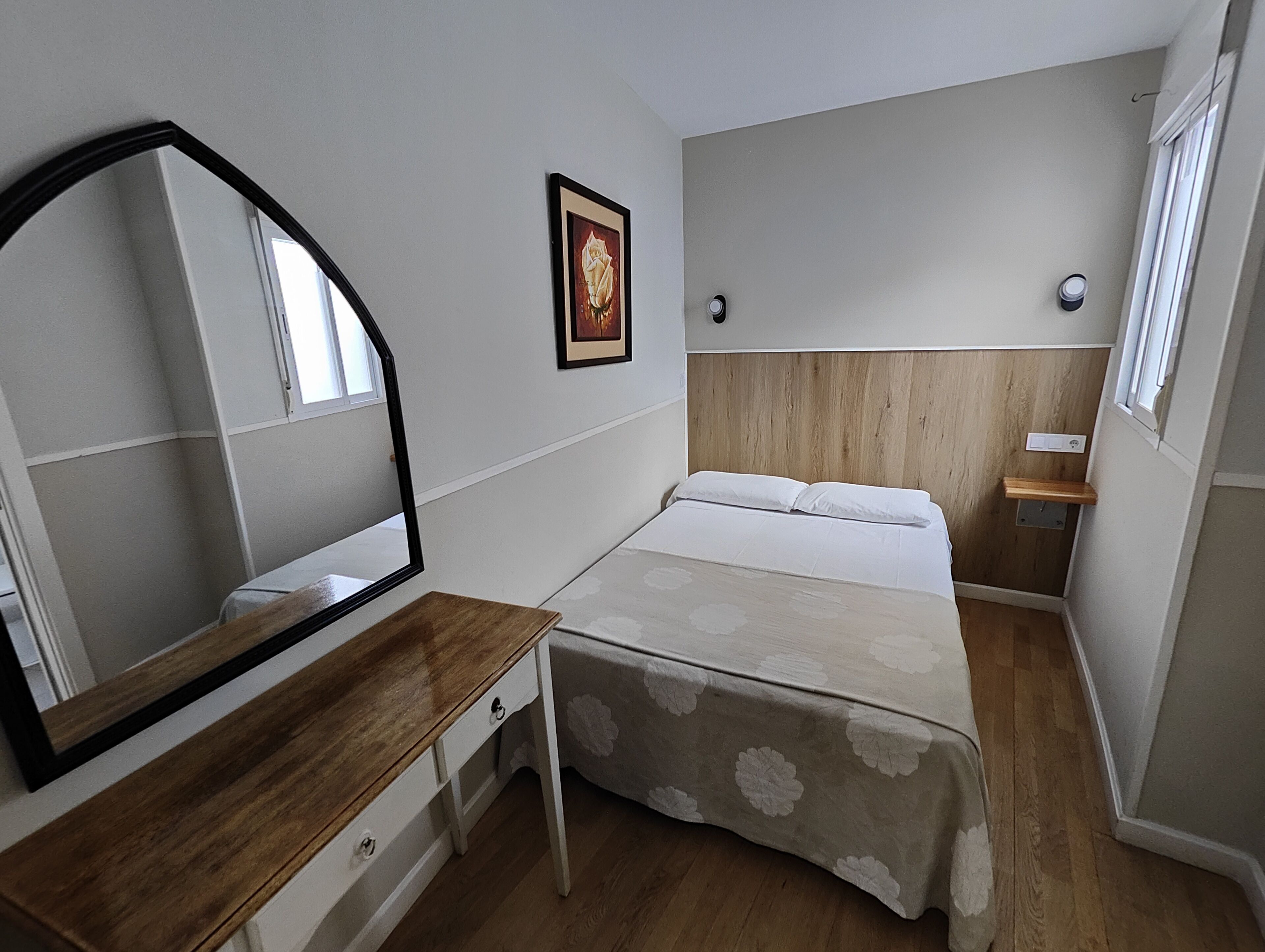 A bedroom with a wooden dresser, a bed with a floral bedspread, a mirror, a framed picture on the wall, and a small shelf.