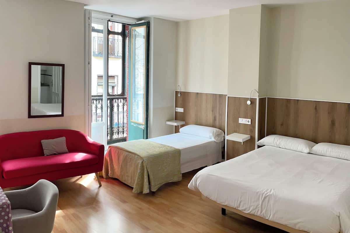Una habitación de hotel con un sofá rojo, una mesita con mantel morado de lunares, una cama con ropa de cama blanca y suelo de madera.