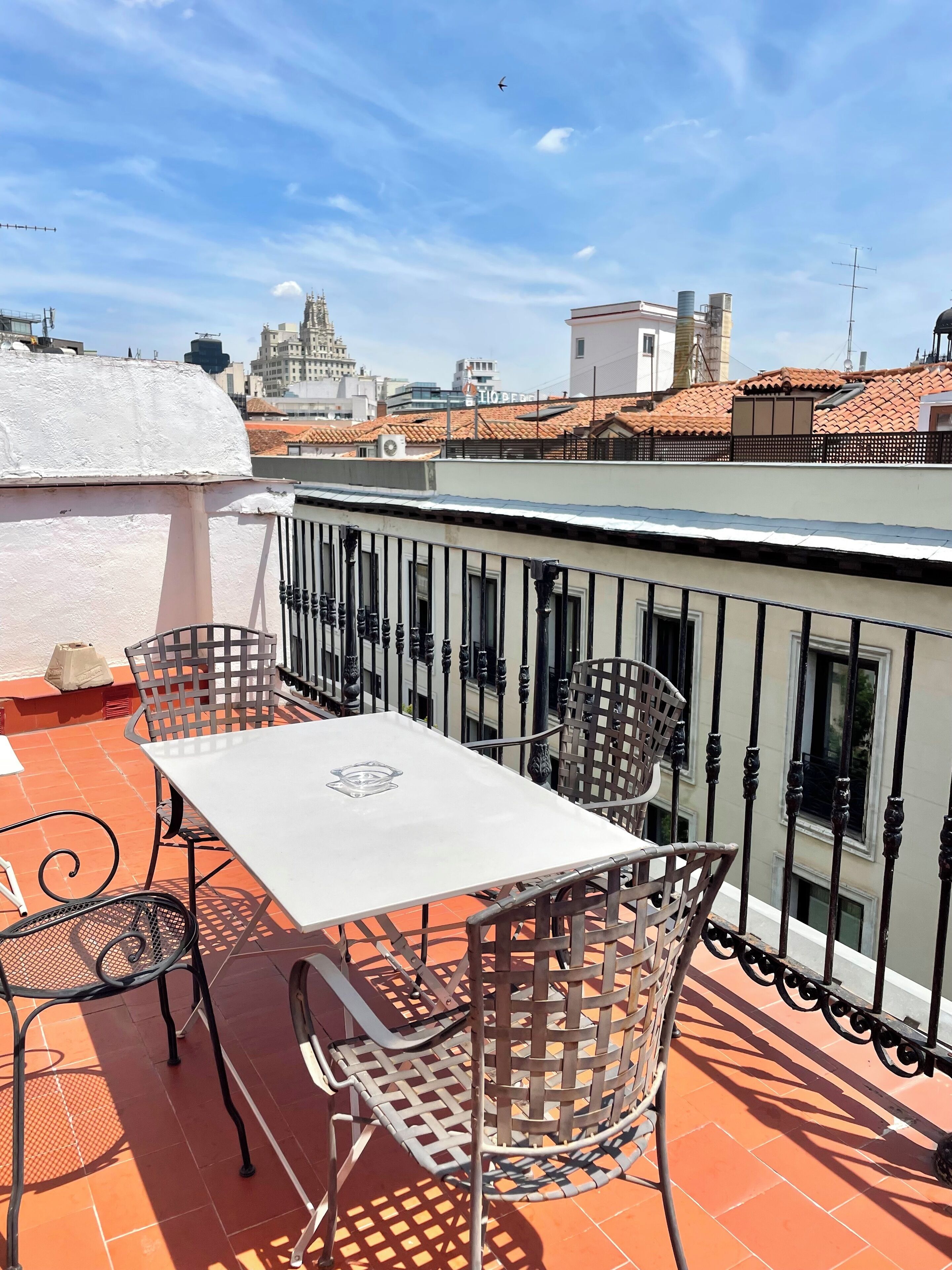 Um terraço no telhado com mesa e cadeiras, vista para a paisagem urbana e um edifício branco com telhado de telhas.