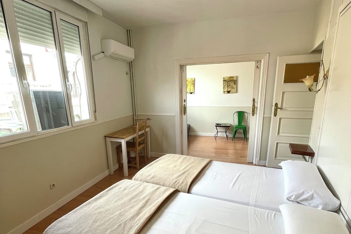 Un dormitorio con dos camas, una mesita pequeña y una silla. Hay una ventana con persianas, una puerta que da a otra habitación y un aire acondicionado instalado en la pared.