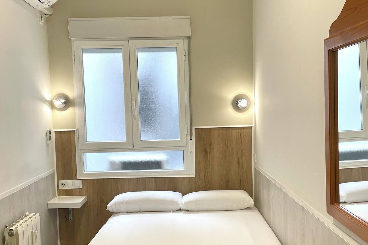 Un dormitorio pequeño y ordenado con una cama individual, una ventana con vidrio esmerilado, una mesita de noche con lámpara y un radiador.