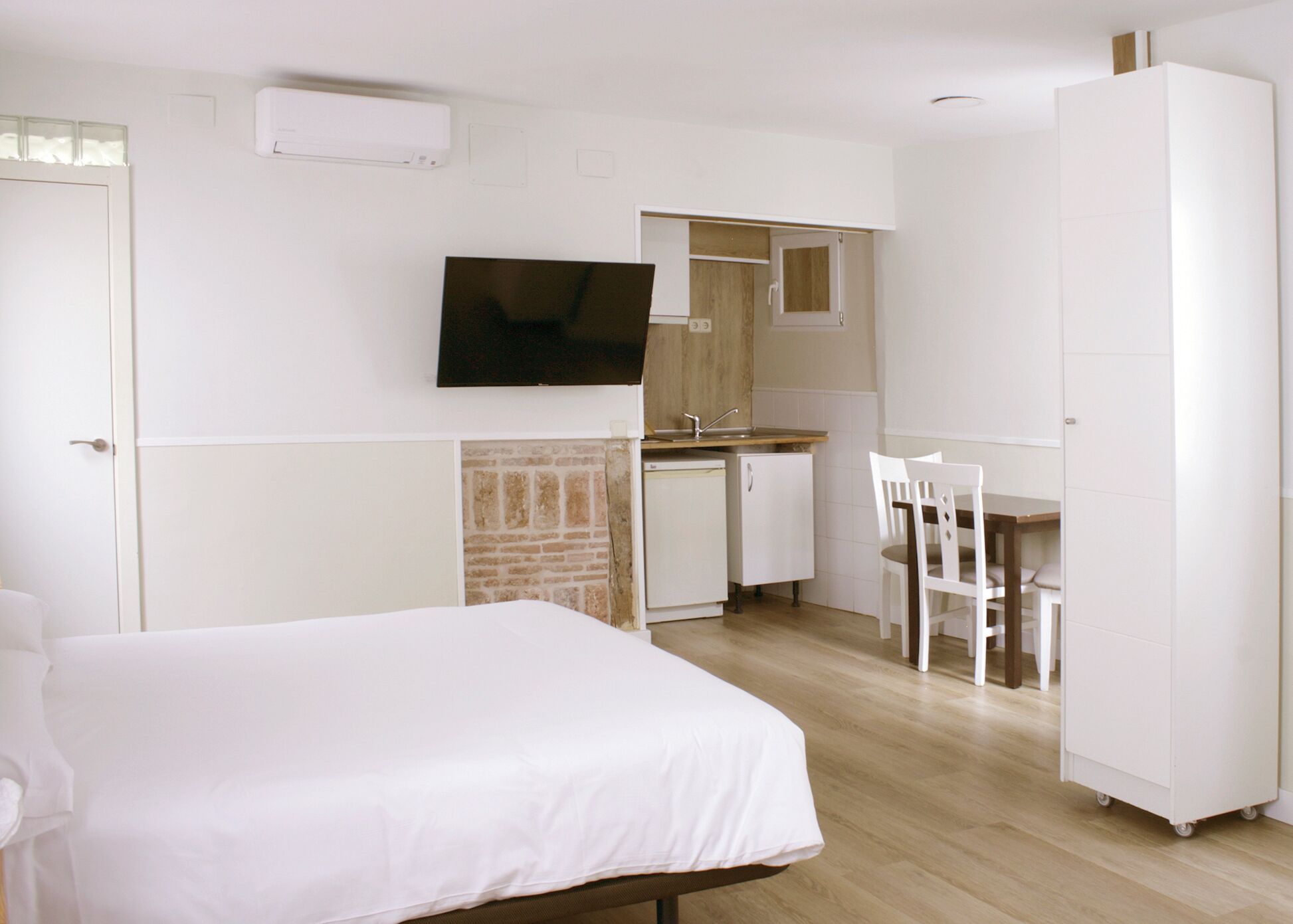 A bedroom with a bed, a TV mounted on the wall, a kitchenette with a sink and cabinets, and a dining area with chairs.