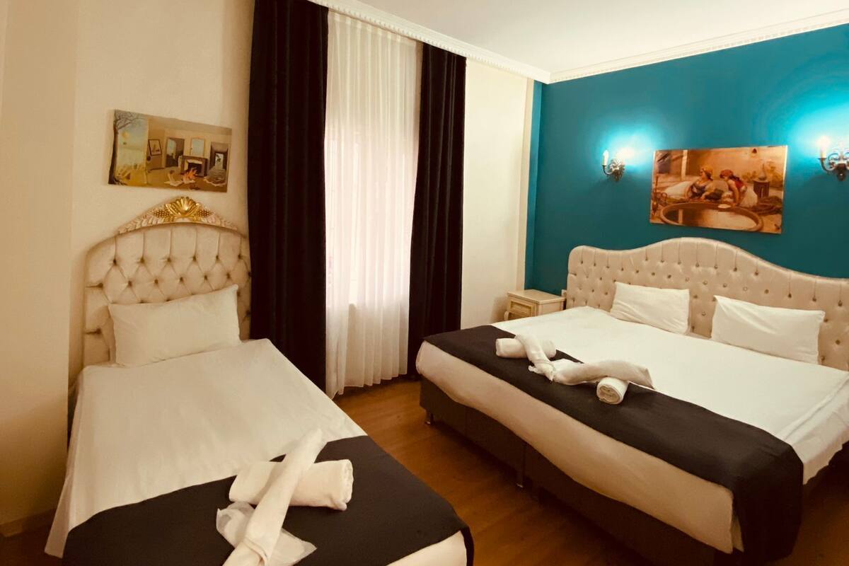 A hotel room with two beds, a large window with curtains, a painting on the wall, and a blue accent wall.