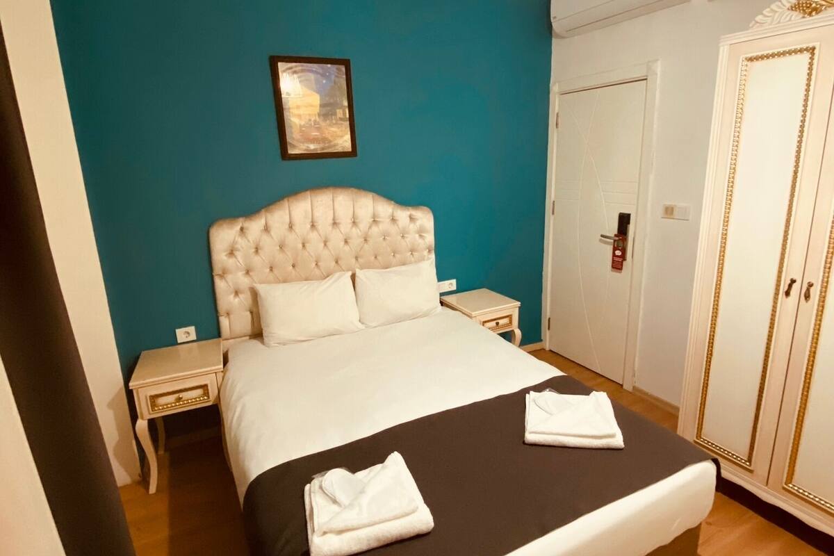 A bedroom with a tufted headboard, a bed with white linens, a black bedspread, two white towels, a nightstand with a lamp, a framed picture on the wall, and a wardrobe with gold detailing.