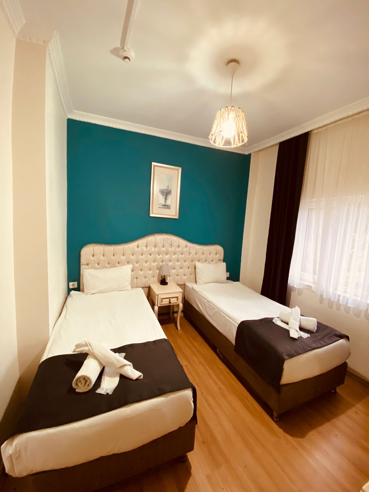 A hotel room with two beds, a teal accent wall, a framed picture, and a hanging light fixture.