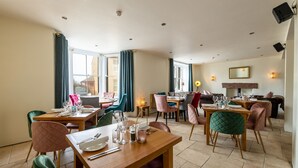 A dining area with multiple tables and chairs, large windows with curtains, and a fireplace. - The Pennington Hotel (Ravenglass)