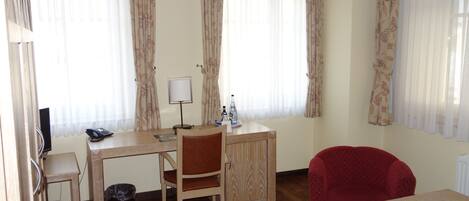 A room with a wooden desk, a chair, a red armchair, a small cabinet, and a trash bin.