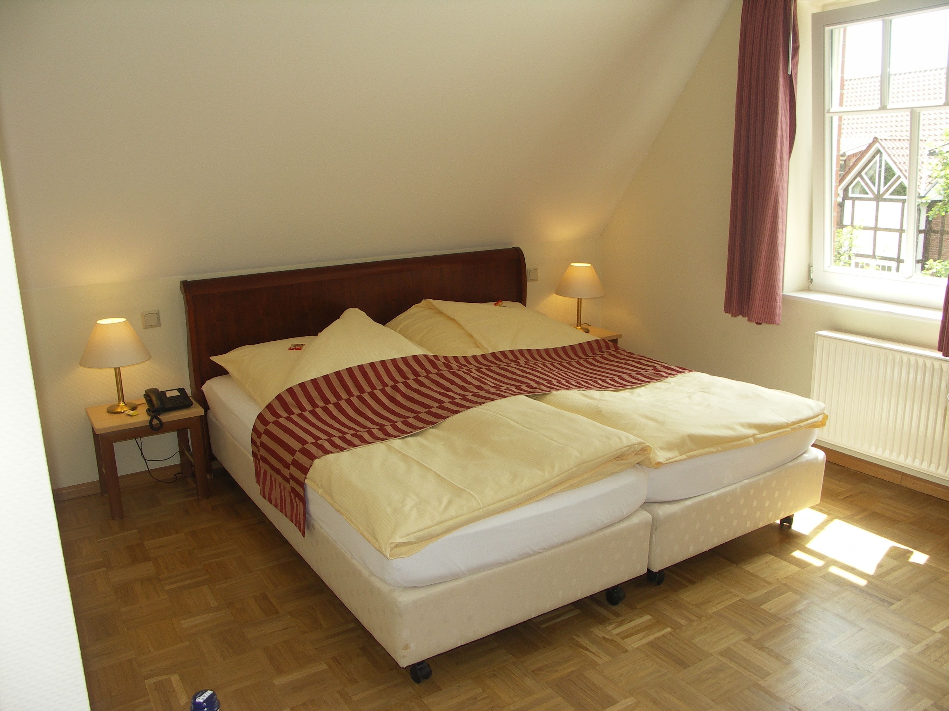 Una cama doble con cabecera de madera, dos mesitas de noche con lámparas y un ventanal con cortinas.