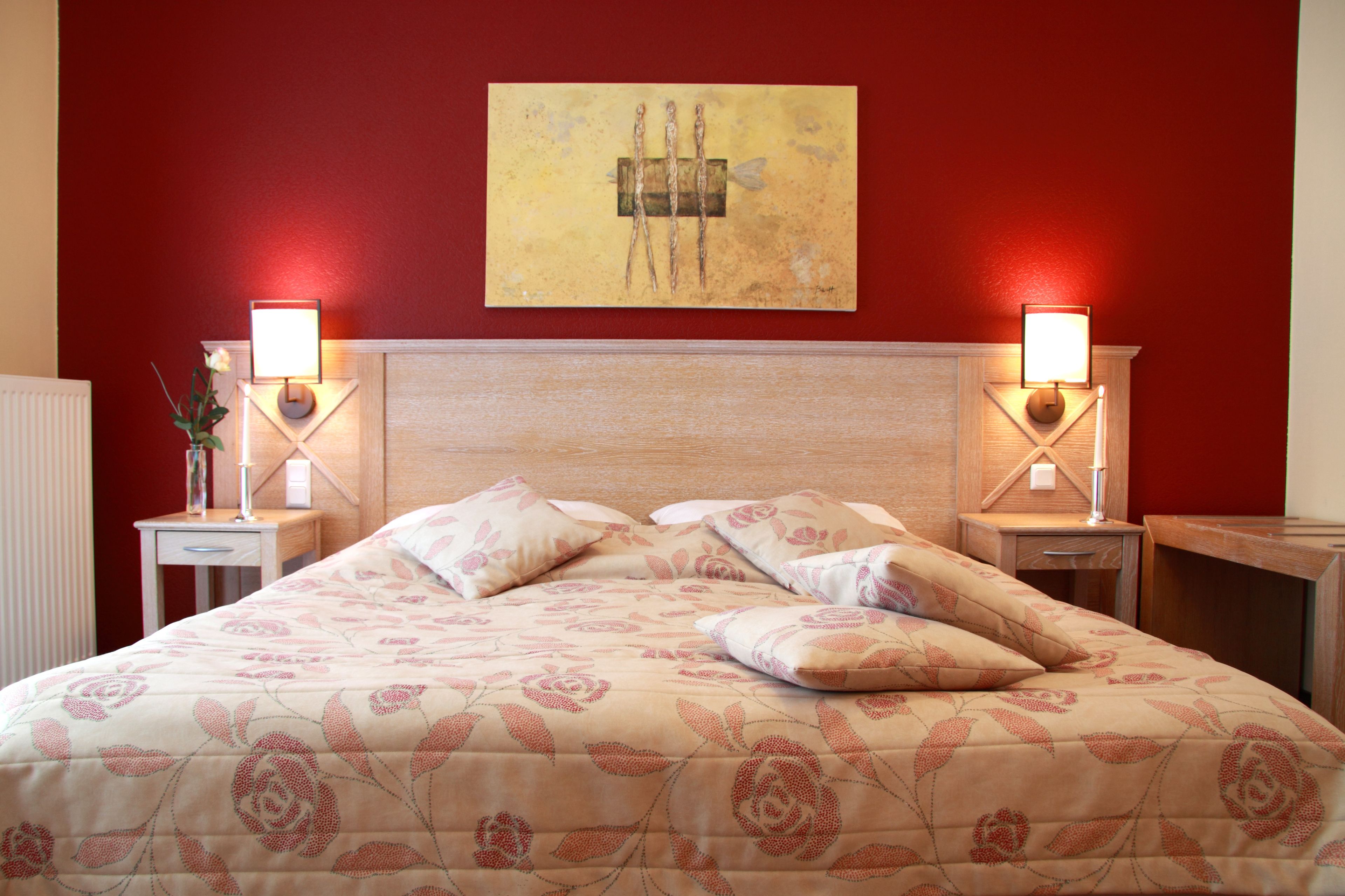 A hotel room with a bed, two bedside tables, a nightstand, a wall-mounted artwork, and a red wall.