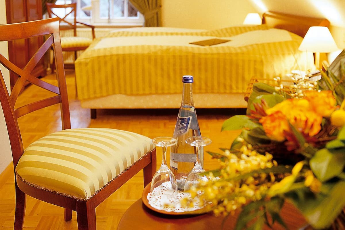 Ein Hotelzimmer mit einem Bett, einem Sessel, einem Tisch mit Flasche und Gläsern sowie einem Fenster mit Vorhängen.