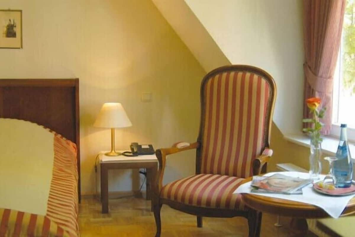 Ein Hotelzimmer mit Bett, Sessel, Schreibtisch mit Lampe, Telefon, einer Vase mit Blume und einem Fenster mit Vorhängen.