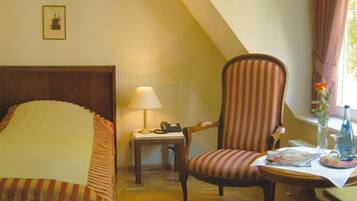 A hotel room with a bed, a chair, a desk with a lamp, a phone, and a vase with a flower.
