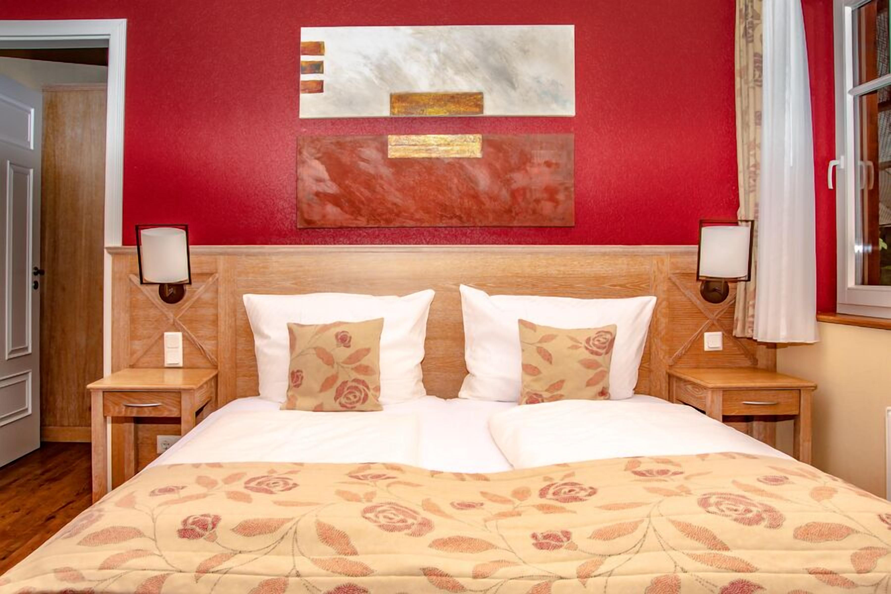 Cama doble con edredón floral, cabecera de madera, dos mesitas de noche con lámparas y una pared roja con arte abstracto.