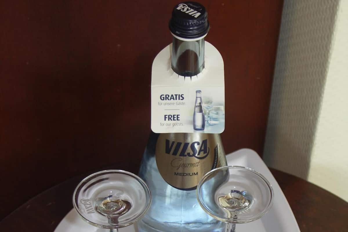 Eine Flasche Vilsal Gourmet Medium Wein mit einem Etikett, das Gästen kostenlosen Wein anbietet, platziert auf einem weißen Tablett mit zwei Weingläsern.