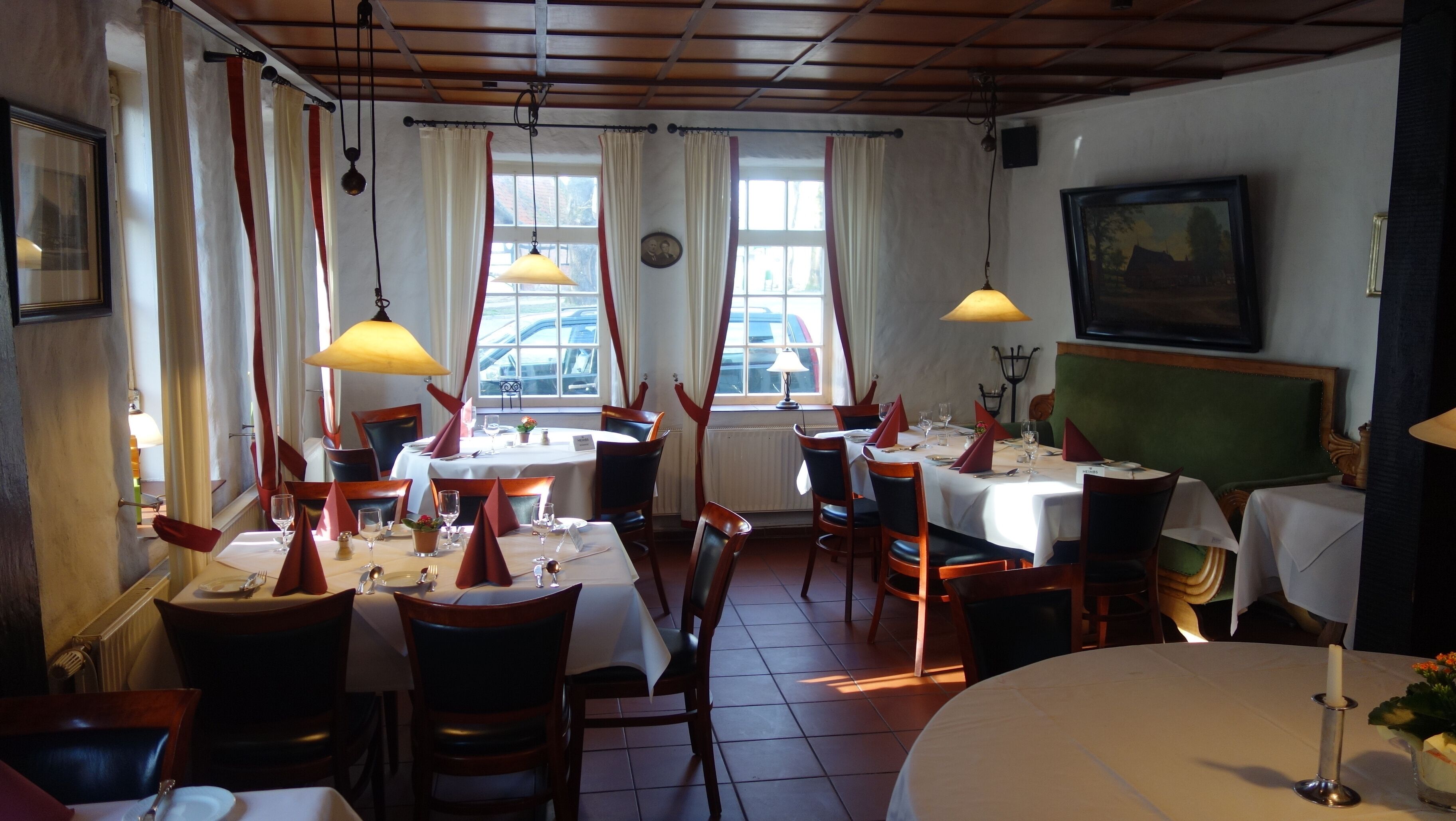A restaurant with multiple tables and chairs, large windows with curtains, and hanging lamps.