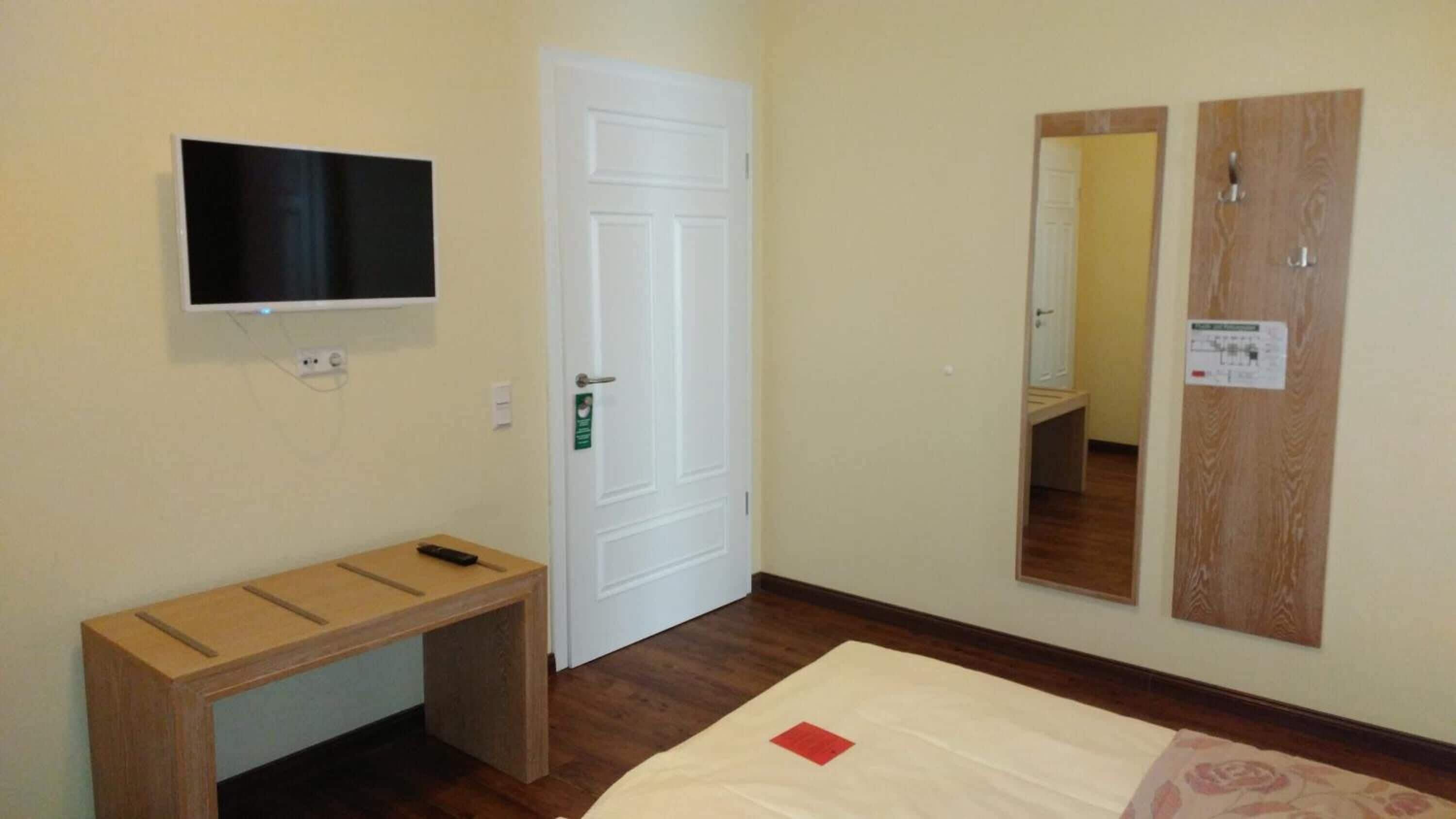 A hotel room with a bed, a TV mounted on the wall, a wooden bench, and a door with a mirror.