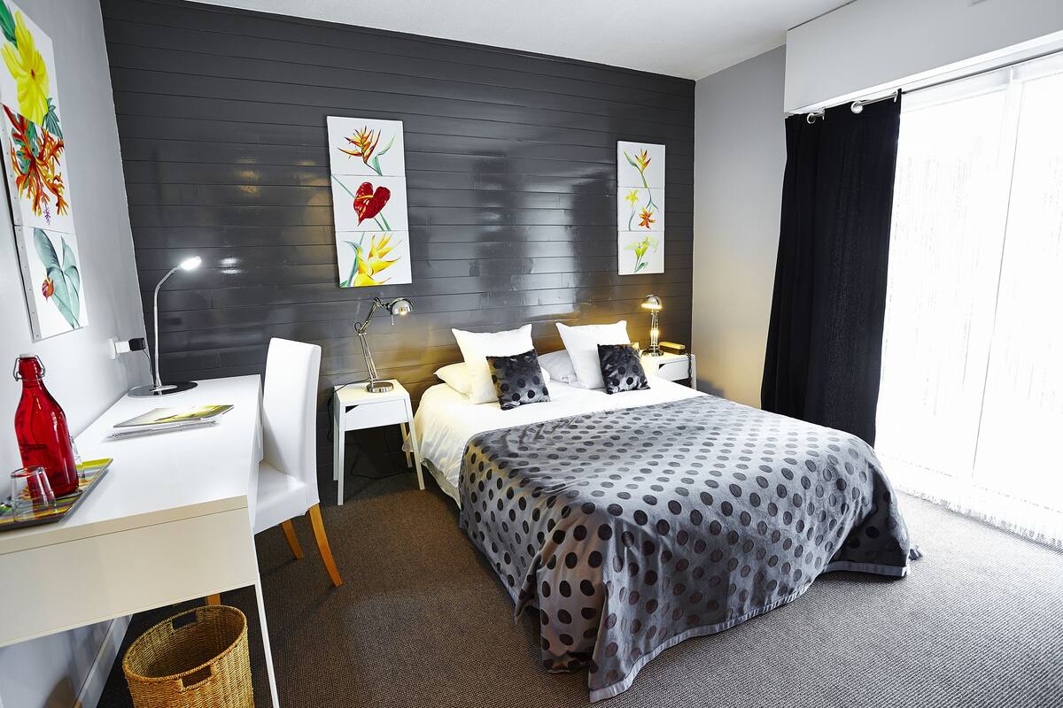 A modern hotel room with a bed, a desk, and a chair. There are two wall-mounted lamps and a window with a black curtain.