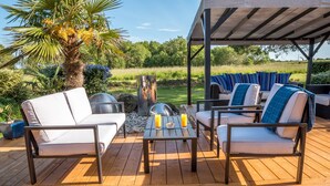 Una terraza al aire libre con piso de madera, dos sofás blancos con cojines azules, una mesa de metal y un área de estar cubierta.