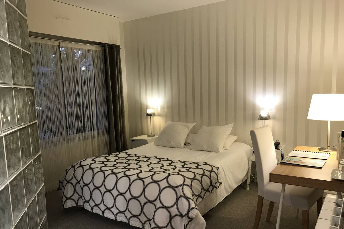 A hotel room with a large bed, two bedside lamps, a desk with a chair, and a window with curtains.
